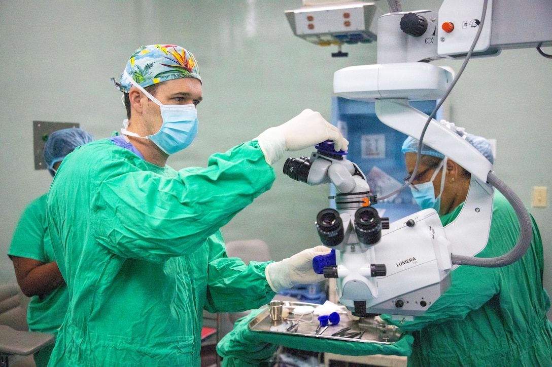A man wearing medical scrubs, a surgical mask and gloves adjusts medical equipment as two other people wearing similar attire stand behind him; a tray of medical instruments and equipment is in the foreground.