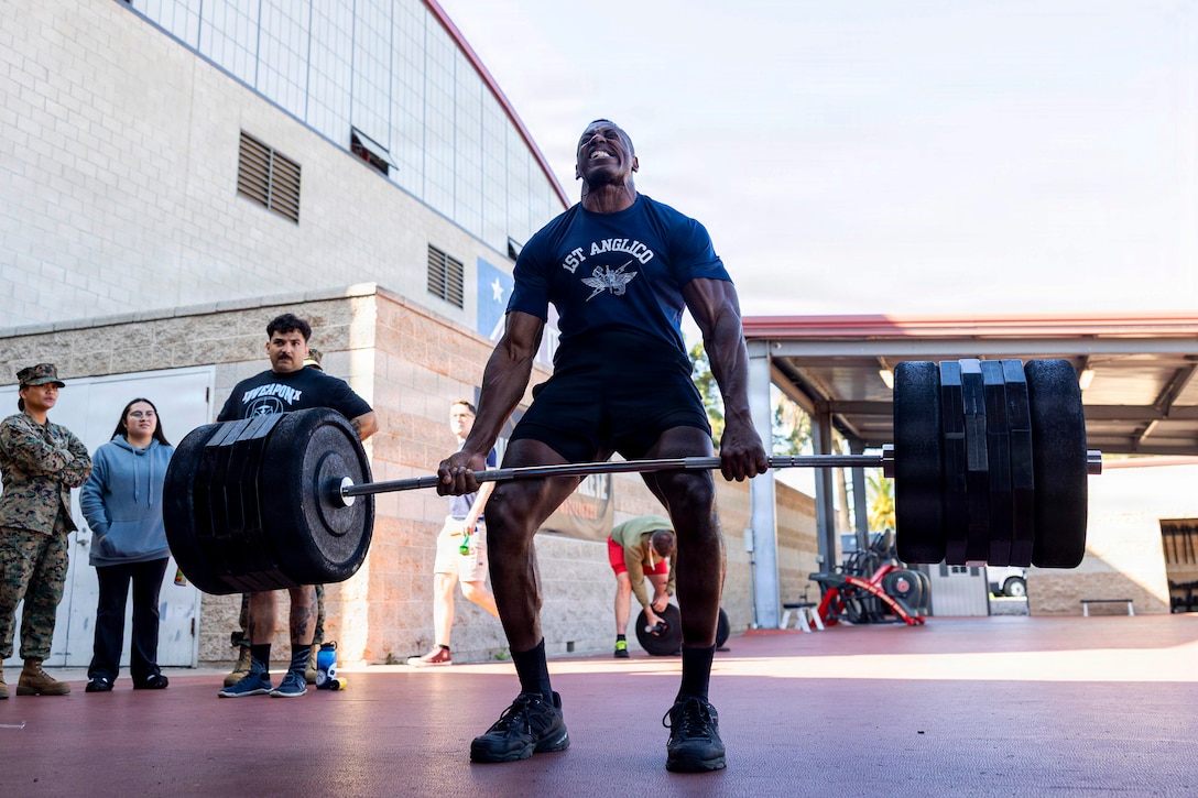 A man wearing athletic attire lifts a barbell with large weights on each end outside near a brick building, as about five people stand nearby and observe.