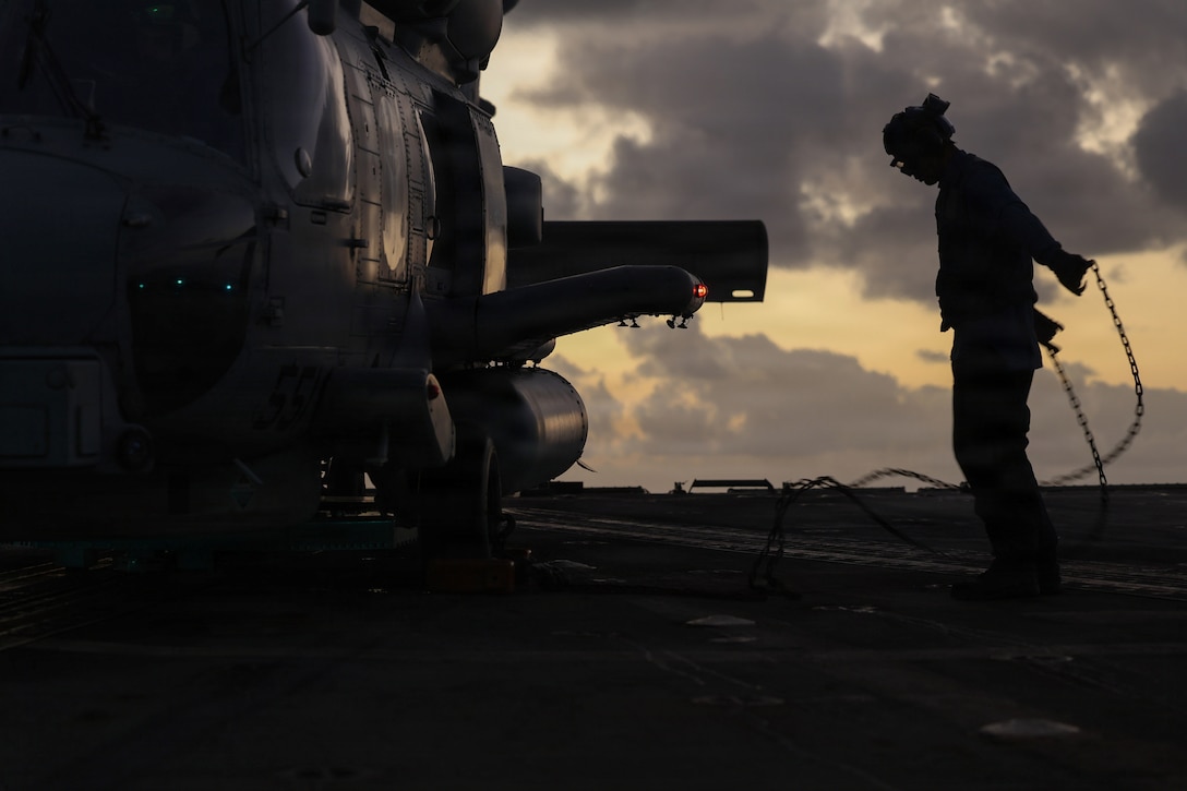 A sailor removes chains from a helicopter aboard a ship as seen in silhouette against a yellowish, cloudy sky.