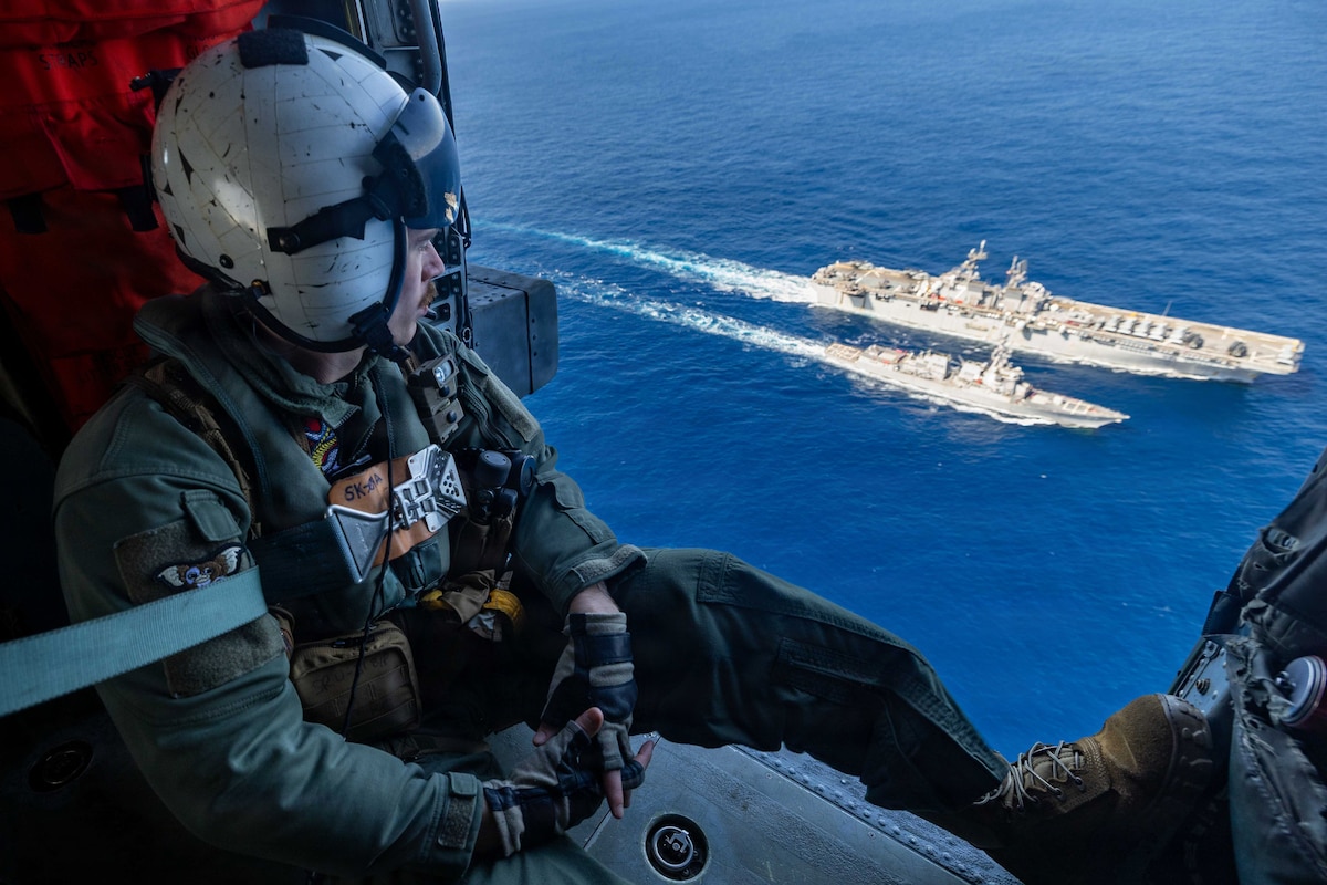 A sailor sits in the open doorway of an airborne helicopter as two ships transit a body of water next to each other.