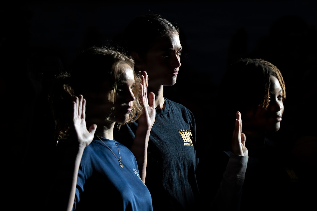 Three people standing together and partly silhouetted against a black background raise their right hands.