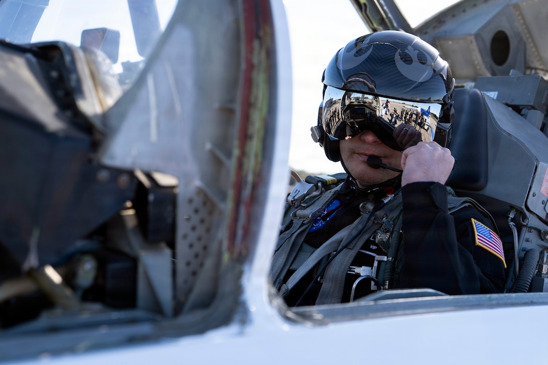 Secretary of War Pete Hegseth adjusts a mouthpiece while wearing a flight helmet and sitting in an aircraft cockpit.