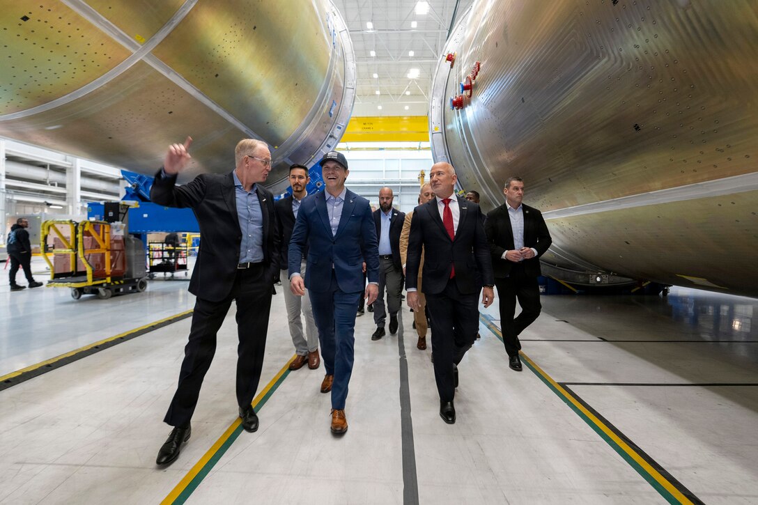 Secretary of War Pete Hegseth walks with several civilians on a factory floor between two large metal objects.
