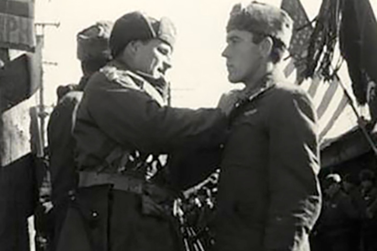 A man pins something to the lapel of another man. Two flags are being presented in the background.
