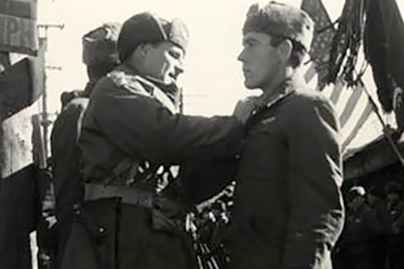 A man pins something to the lapel of another man. Two flags are being presented in the background.