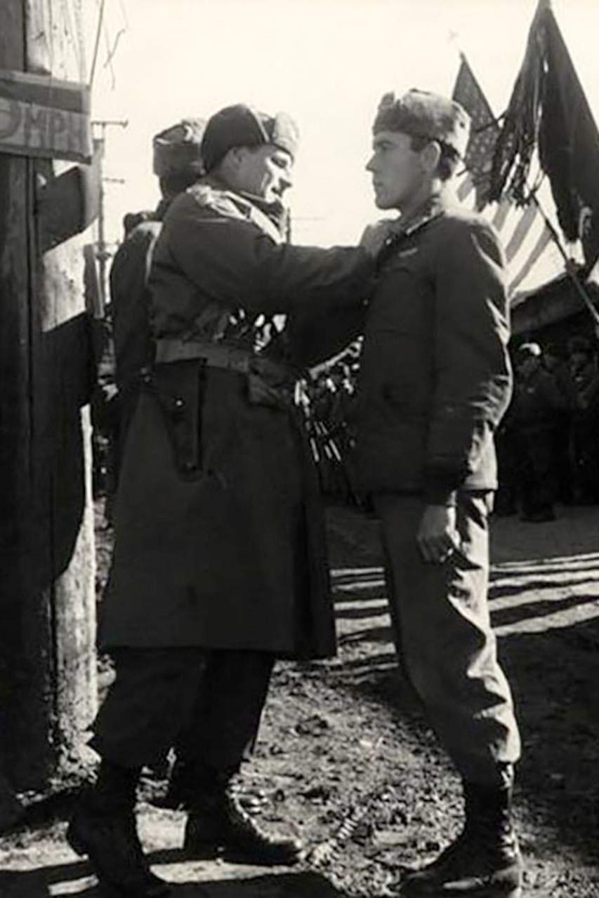 A man pins something to the lapel of another man. Two flags are being presented in the background.
