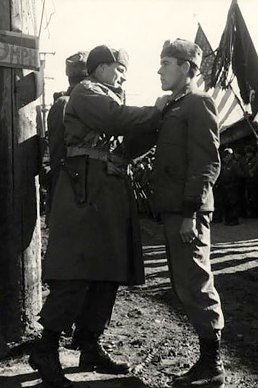 A man pins something to the lapel of another man. Two flags are being presented in the background.