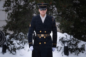 An Army Soldier wearing ceremonial winter coat and white gloves is standing at attention in front of a snowy scene in the background.