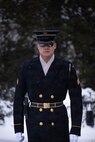 An Army Soldier in ceremonial winter coat with white gloves is standing at attention and there is snow on the ground in the background.