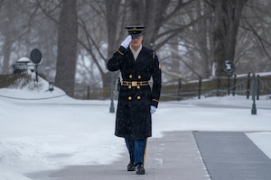 An Army Soldier wearing ceremonial winter coat and white gloves is saluting while walking near the black mat used by Tomb Guards at the Tomb of the Unknown Soldier, and there is snow on the ground in the background.