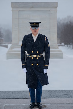 An Army Soldier wearing a ceremonial winter coat, sunglasses, hat and white gloves is facing the camera while standing at attention in front of the white marble-clad Tomb of the Unknown Soldier with snow on the ground.