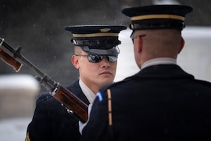 Two Army Soldiers earing ceremonial uniforms with sunglasses and hats on are facing each other as one is holding a ceremonial rifle at a 45 degree angle in front of his chest.