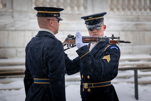An Army Soldier on the right is holding a ceremonial rifle up close to his face as he inspects it while another Soldier on the left watches.  Both are wearing ceremonial coats with white gloves, and there is snow on the ground in the background.