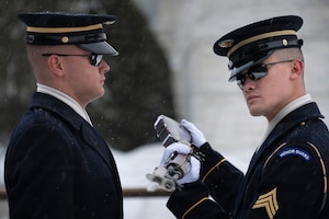 An Army Soldier on the right is inspecting a ceremonial rifle for another Soldier who is on the left. Both are wearing ceremonial uniforms, and there is snow on the ground in the background.