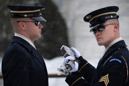 An Army Soldier on the right is inspecting a ceremonial rifle for another Soldier who is on the left. Both are wearing ceremonial uniforms, and there is snow on the ground in the background.