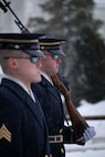 Two Army Soldiers wearing ceremonial uniforms with white gloves and sunglasses are standing next to each other as one is carrying a ceremonial rifle.