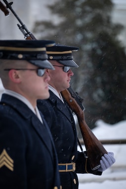 Two Army Soldiers wearing ceremonial uniforms with white gloves and sunglasses are standing next to each other as one is carrying a ceremonial rifle.