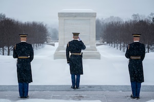 Three U.S. Army Soldiers wearing ceremonial uniforms are standing in a triangle formation facing away from the camera as the middle soldier salutes in the direction of the white marble-clad Tomb of the Unknown Soldier with snow on the ground in the background.