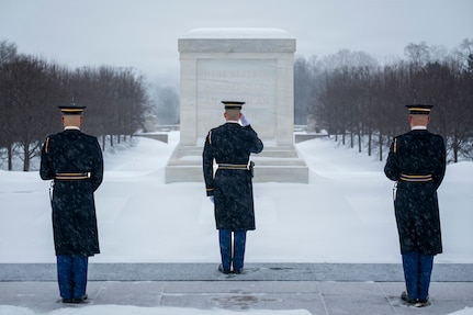 Three U.S. Army Soldiers wearing ceremonial uniforms are standing in a triangle formation facing away from the camera as the middle soldier salutes in the direction of the white marble-clad Tomb of the Unknown Soldier with snow on the ground in the background.