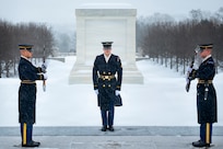 Three Army Soldiers wearing ceremonial uniforms, two holding ceremonial rifles in front of their chests facing each other while the other faces the camera, are standing in front of the white marble Tomb of the Unknown Soldier with snow on the ground in the background.