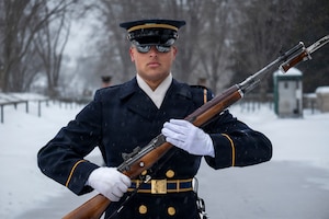 An Army Soldier wearing a ceremonial uniform with white gloves and white scarf is holding a brown ceremonial rifle at a 45 degree angle in front of his chest as he marches and the ground