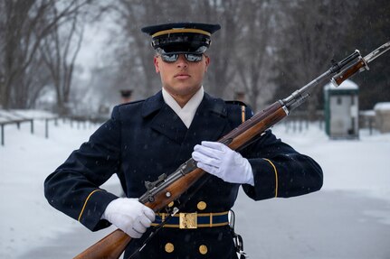 An Army Soldier wearing a ceremonial uniform with white gloves and white scarf is holding a brown ceremonial rifle at a 45 degree angle in front of his chest as he marches and the ground