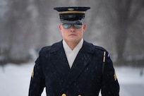 An Army Soldier wearing a ceremonial uniform with coat and white scarf has on sunglasses while standing in a snowy scene.