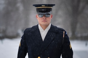 An Army Soldier wearing a ceremonial uniform with coat and white scarf has on sunglasses while standing in a snowy scene.