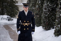 An Army Soldier dressed in a dark blue ceremonial winter coat with hat and gloves is marching along a path with snow along the ground and tall, green bushes in the background.