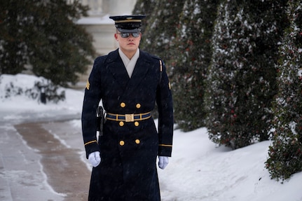 An Army Soldier dressed in a dark blue ceremonial winter coat with hat and gloves is marching along a path with snow along the ground and tall, green bushes in the background.