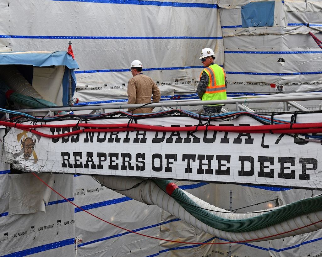 Two people in hard hats walk across a metal bridge leading to a ship.