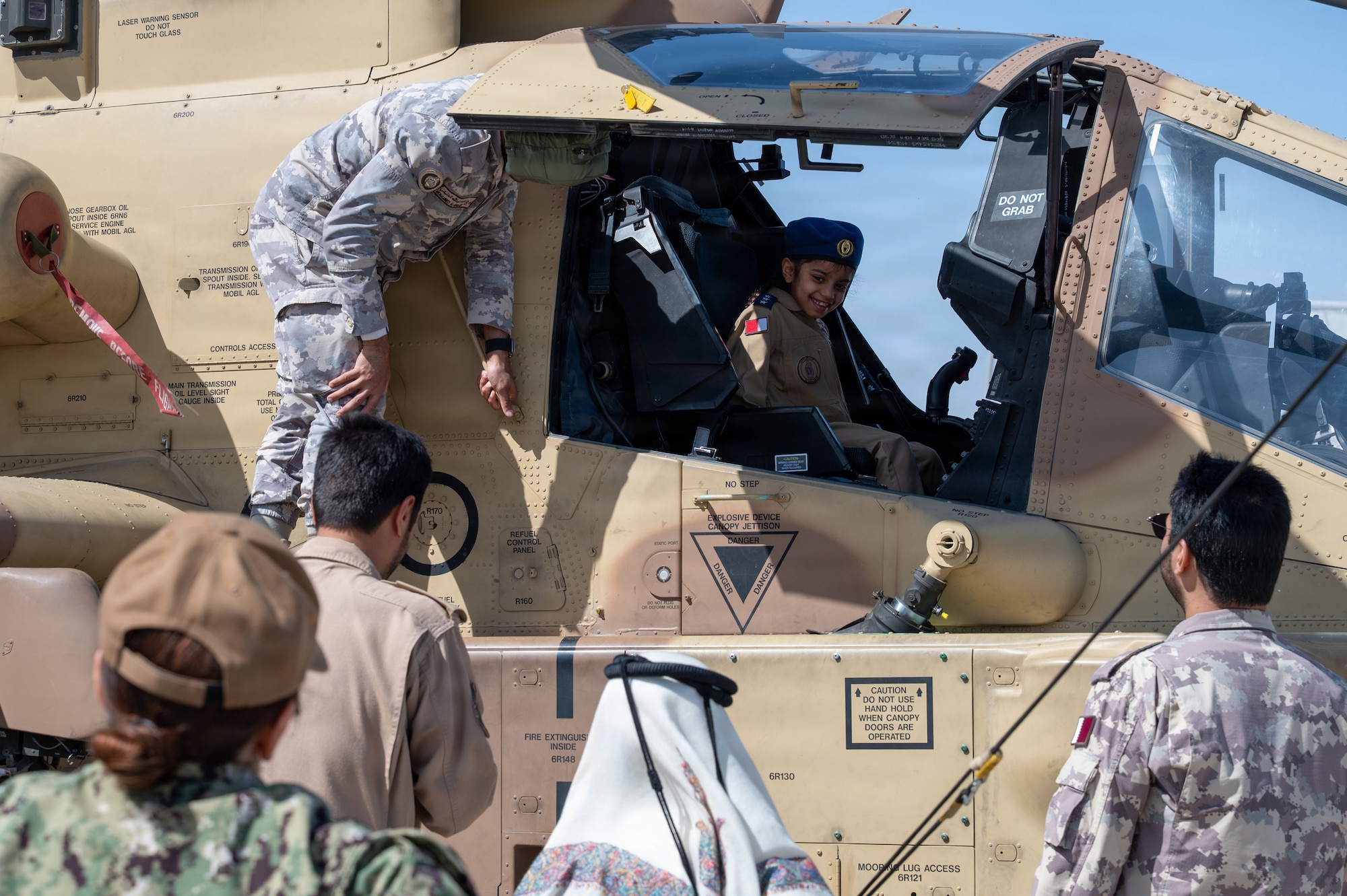 A child sits in the cockpit of a Qatar Emiri Air Force AH-64E helicopter while a QEAF member stands on the aircraft monitoring the child. Others are in the foreground watching from the ground.