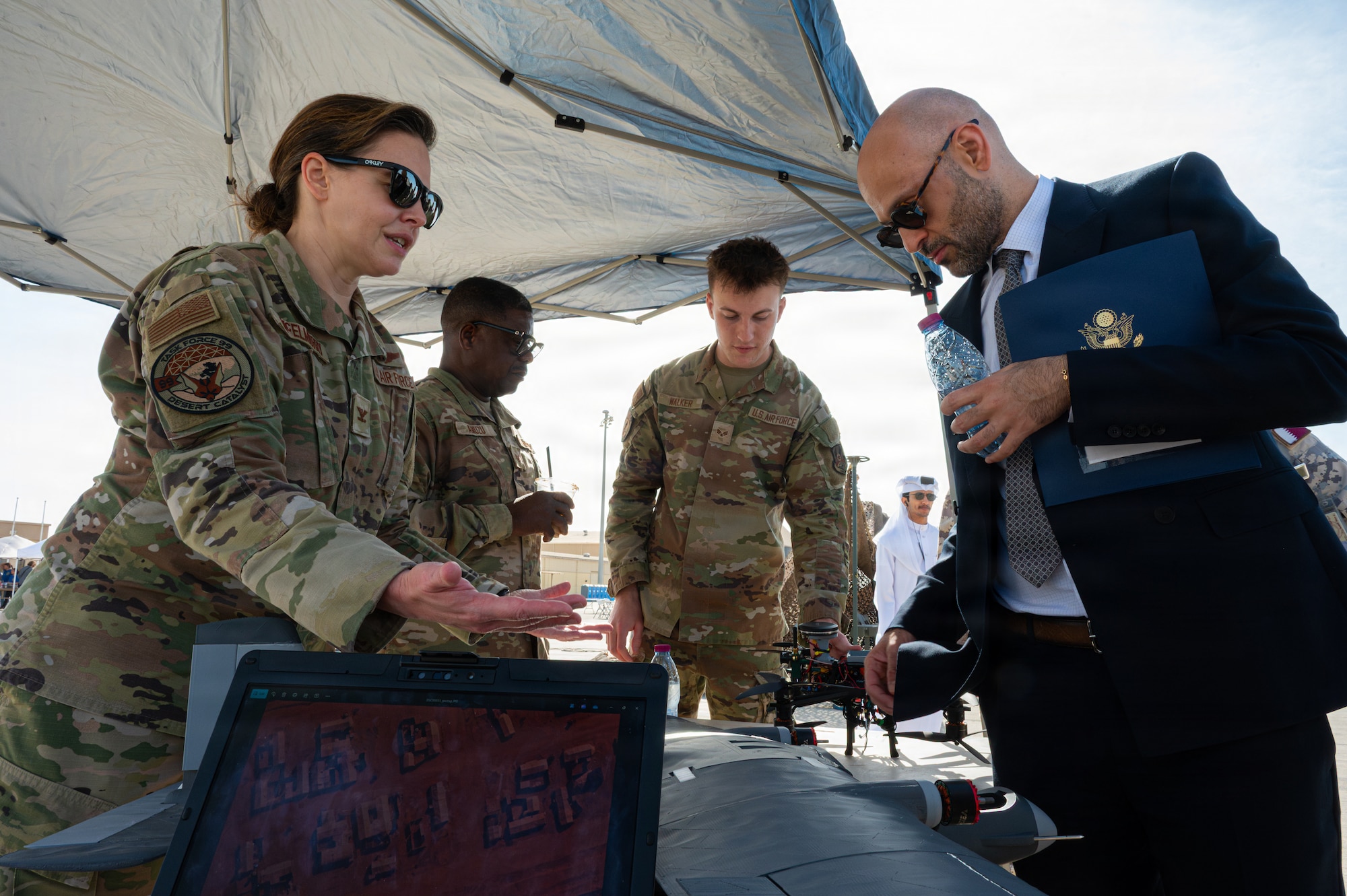An Airman gestures to an unidentifiable object on a table under a canopy while Mo Barghouty looks on. Other Airmen are in the background showing someone a drone.