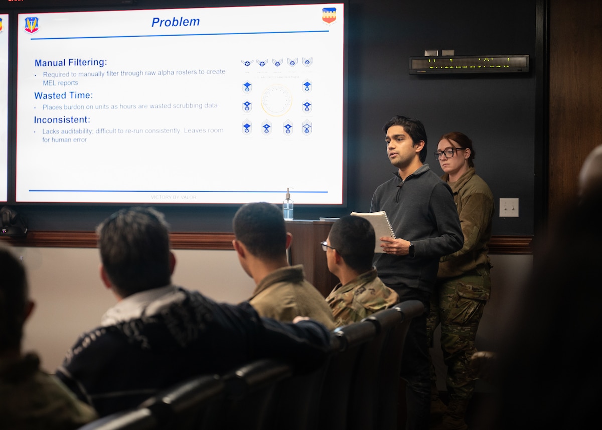 A man in civilian clothes and a woman in an Air Force uniform stand at a podium in the front of a room. There is a slideshow on the projector, and there are sveeral people sitting in the audience, listening to the presentation.