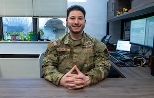 U.S. Airman sits at a desk smiling with hands folded.