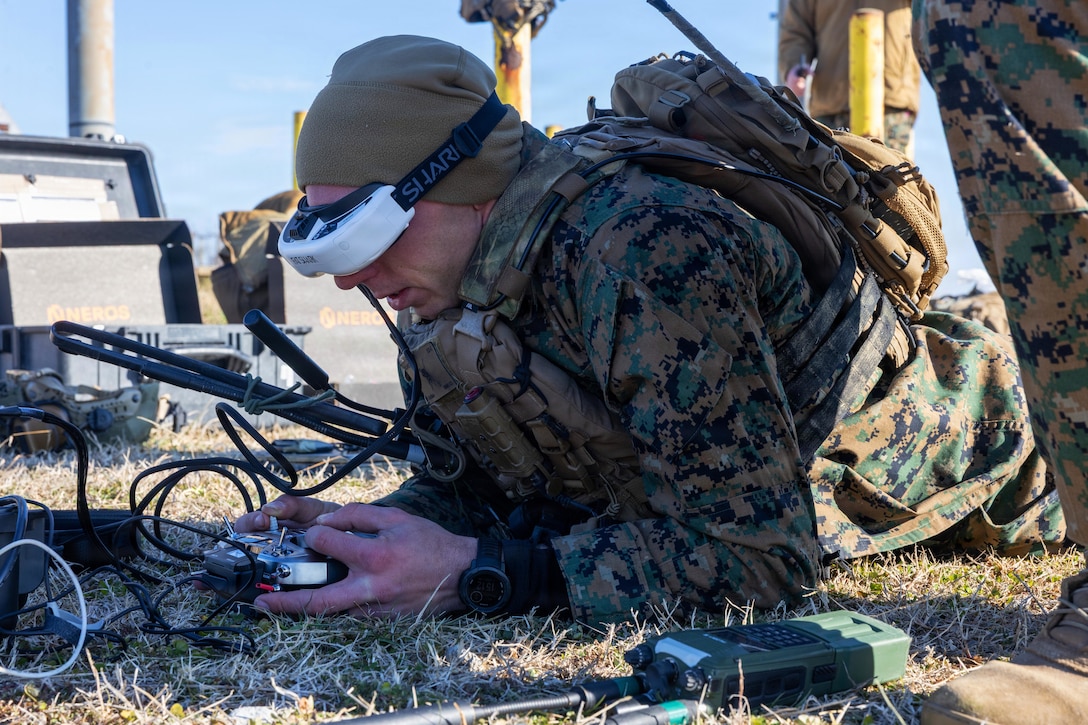 A man lying in a grassy area and wearing a camouflage military uniform, head cover and virtual headset holds a remote control with wires attached to the headset; another person stands to his right and virtual equipment is in the background.