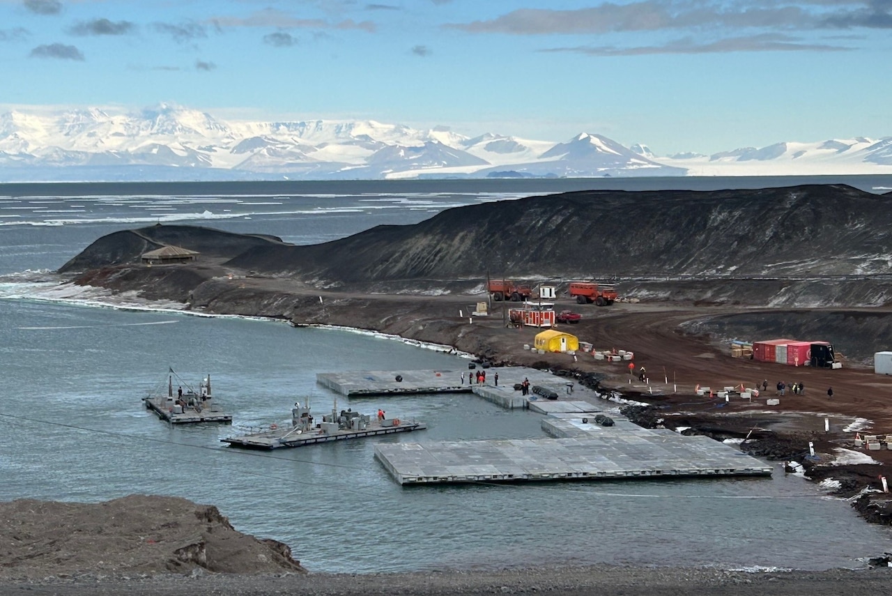 People assemble floating metal pieces into a temporary bridge off a rocky shoreline. There are snow-covered mountains in the background.