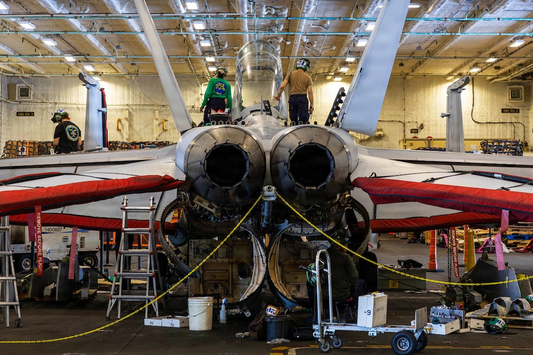 Three rear-facing people in casual attire stand on top of a military jet inside a hangar bay.