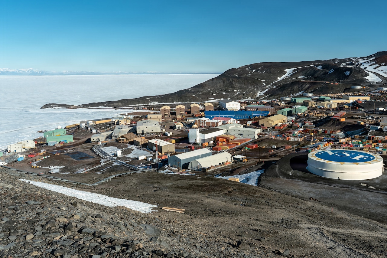 A landscape view of a coastal town in Antarctica with buildings and cargo containers. There are hills with snow in the background, and the water is frozen.