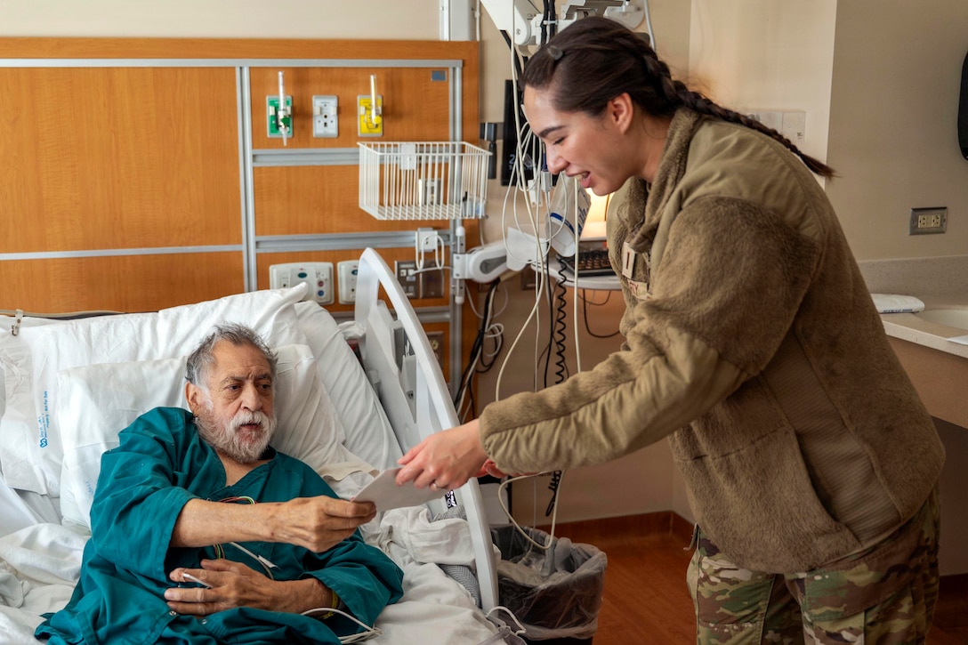 A woman wearing camouflage military pants and a tan military jacket stands and gives a card to a man lying in a hospital bed wearing a green hospital gown. Hospital equipment is in the background.