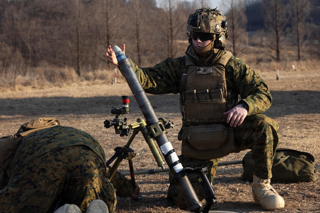 A person wearing a camouflage military uniform, vest and helmet kneels in a rocky area while placing a mortar into the opening of a weapon; another person wearing similar attire crouches nearby; trees and shrubbery are in the background.
