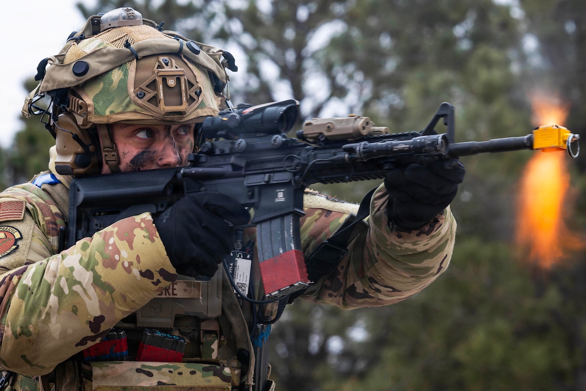 A man wearing a camouflage military uniform, helmet and gloves holds a weapon and fires outside in a wooded area; orange flares emit from the weapon's nozzle.