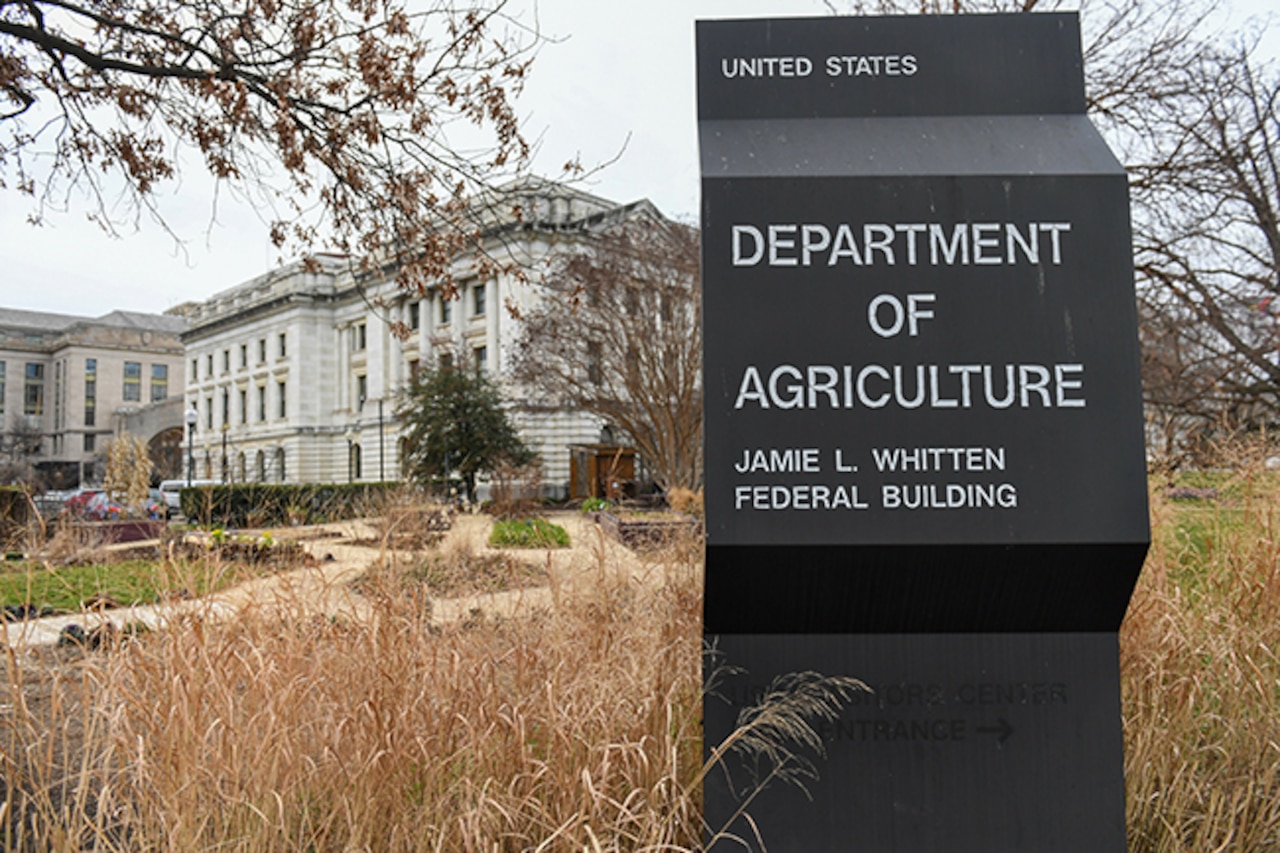 A sign in tall grass reads, "United States Department of Agriculture Jamie L. Whitten Federal Building." A building is in the background.