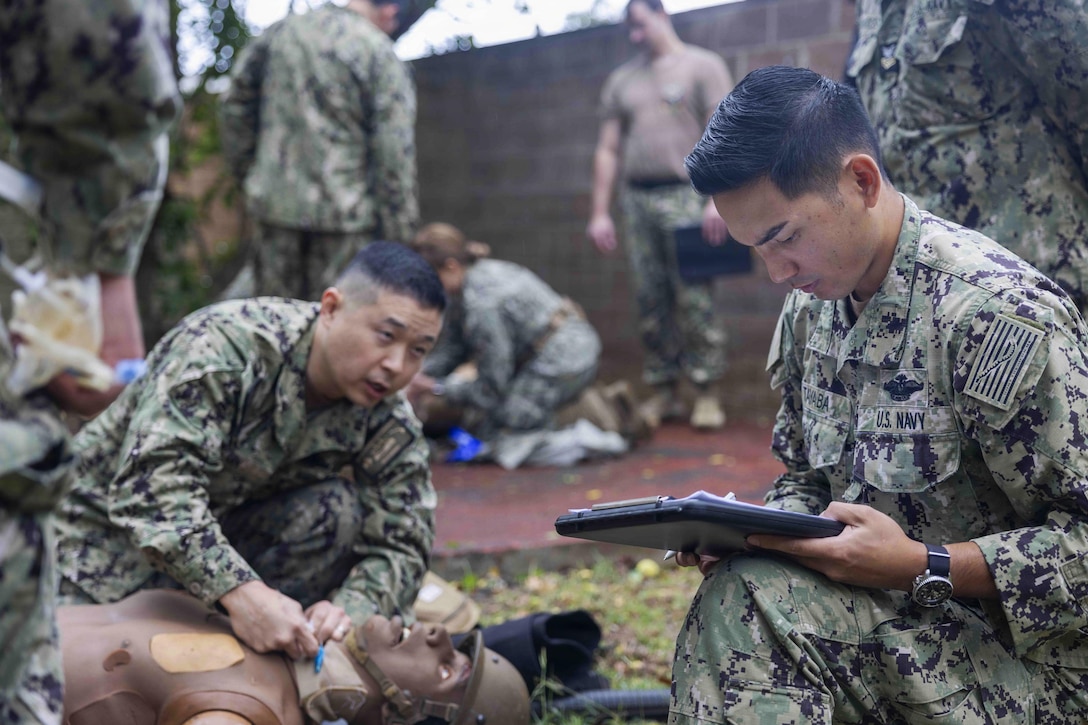 Two people wearing camouflage military uniforms kneel on the ground; one looks upward while practicing on a mannequin, and the other looks down at a clipboard. About five people in similar attire are in the background, engaging in similar activities.