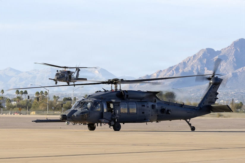 A helicopter hovers over a tarmac while another hovers in the background. There are mountains and a city in the distance.