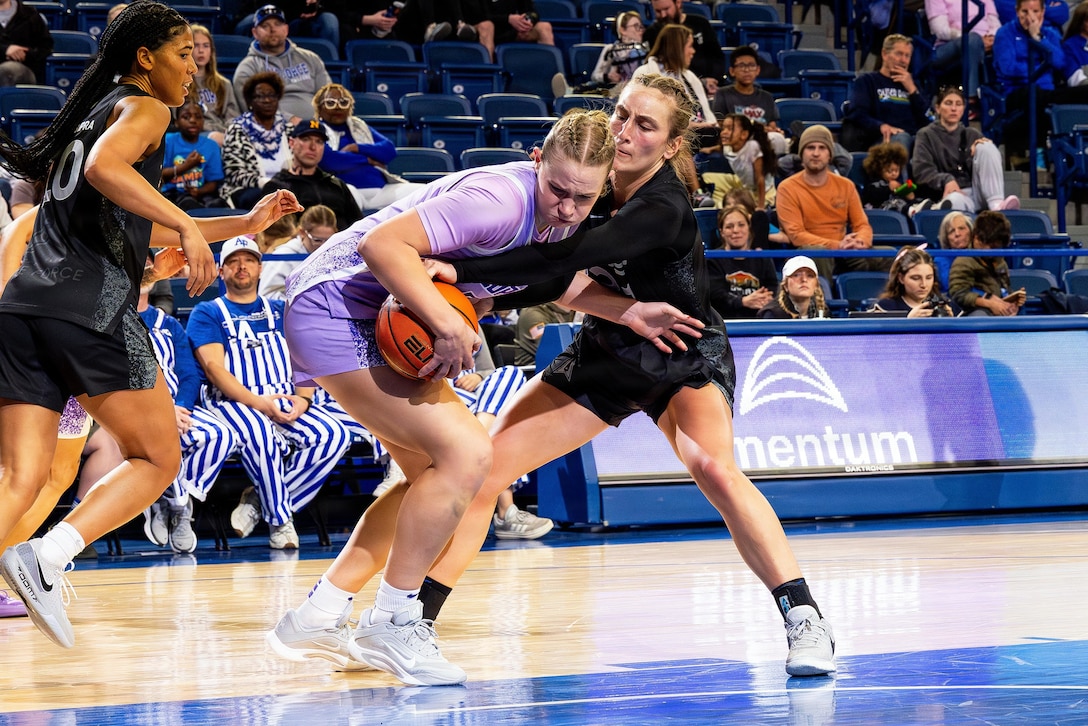 Three women wearing athletic attire play basketball inside a gymnasium as dozens of spectators watch from the stands.