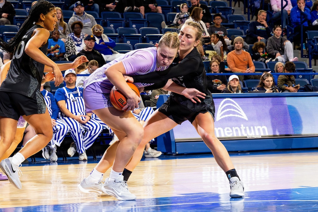 Three women wearing athletic attire play basketball inside a gymnasium as dozens of spectators watch from the stands.