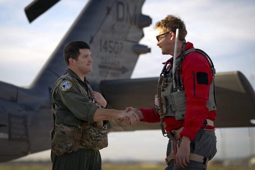 Two men in flight suits shake hands in front of an aircraft.