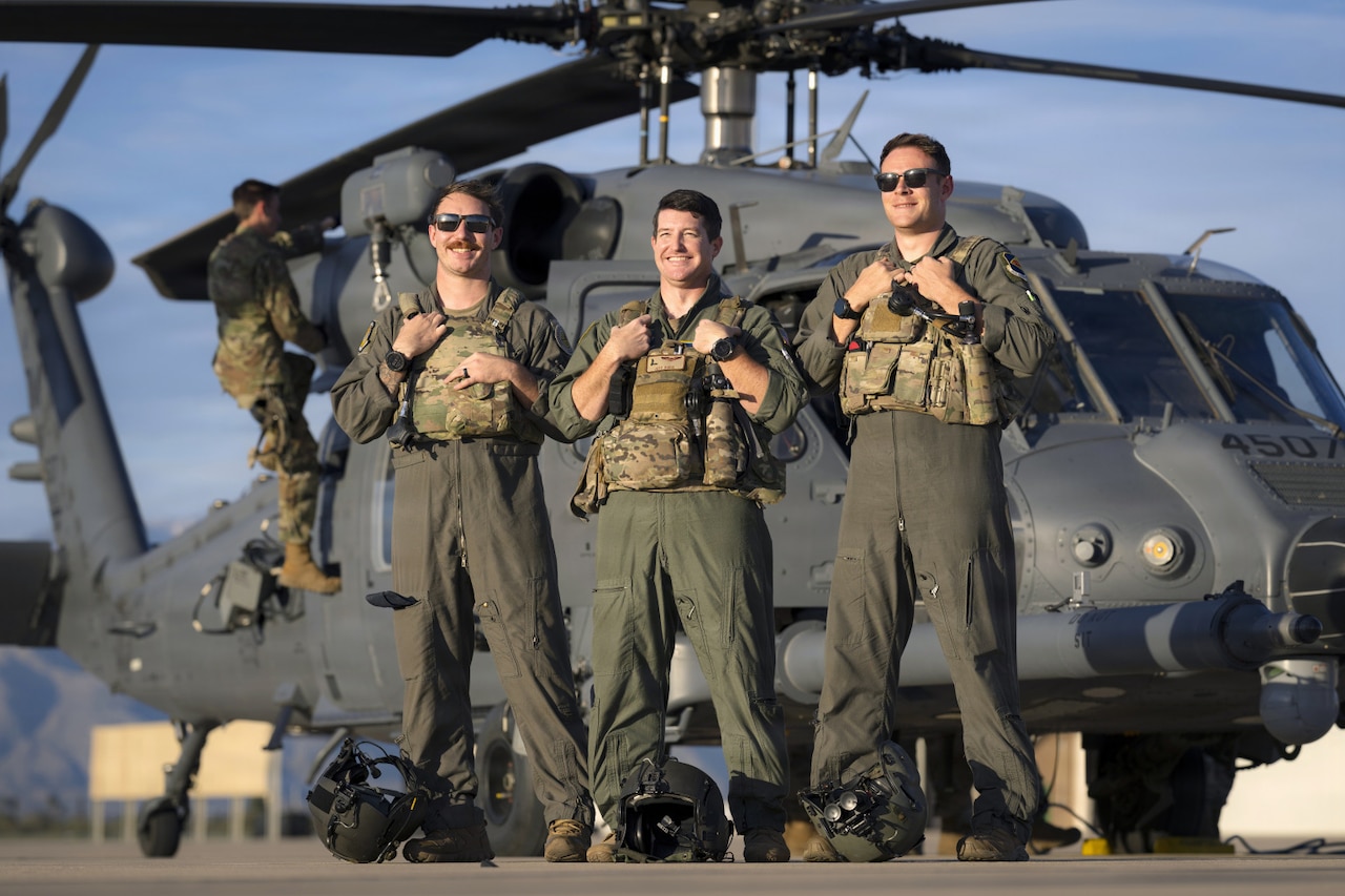 Three men wearing flight suits pose for a photo in front of a helicopter. A man in similar attire is climbing up the side of the helicopter in the background.