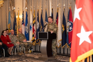 A man in uniform stands at podium facing an audience with a bunch of flags in the room